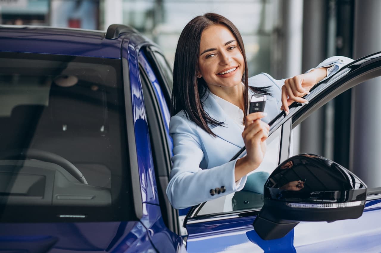 Mulher sorridente de cabelos castanhos channel, segurando a chave de um veículo azul no qual ela está apoiada e segurando a porta de motorista.