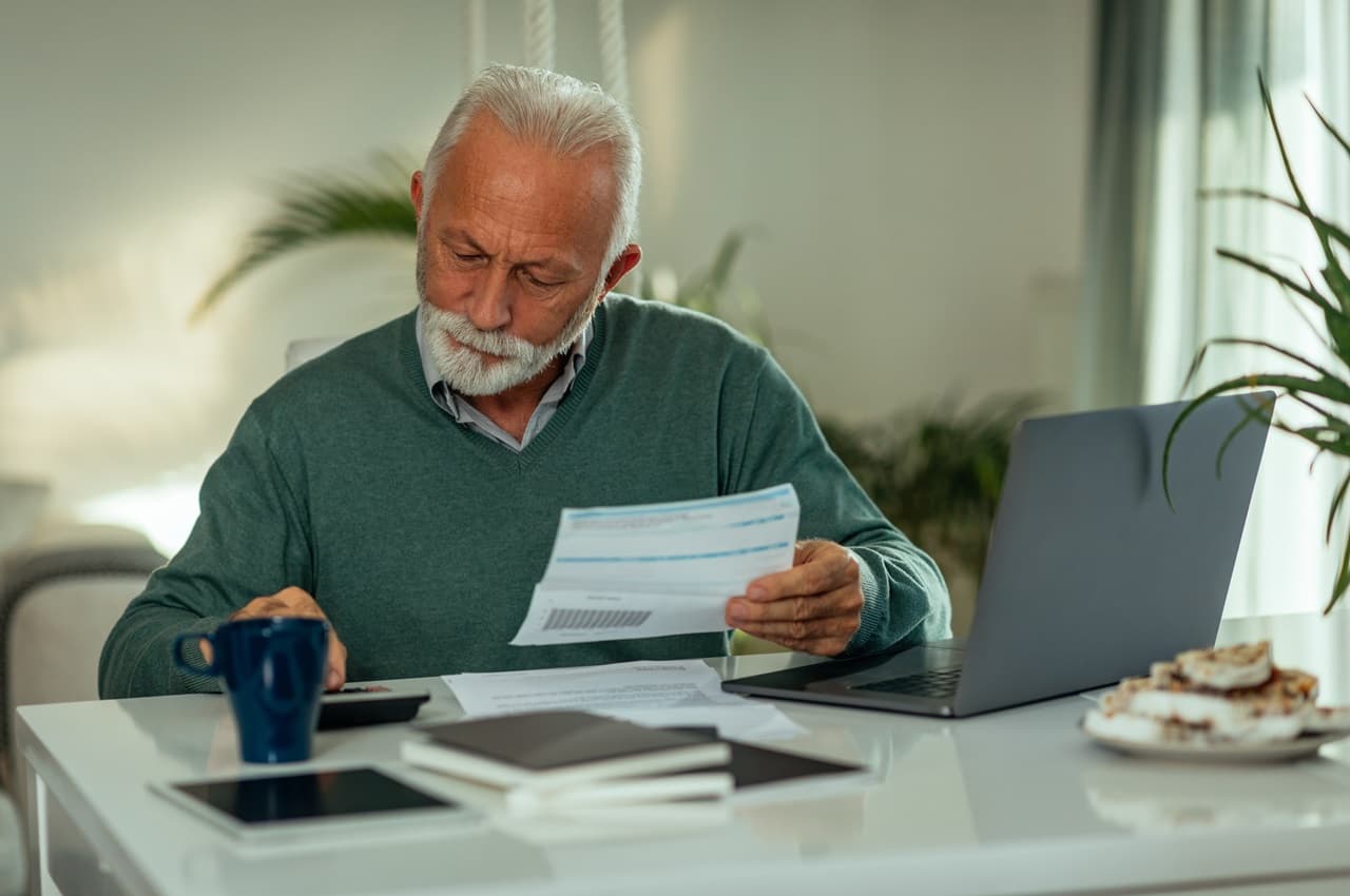 Homem sênior de cabelos curtos brancos segurando parcelas do financiamento e usando calculadora, computador e anotações.