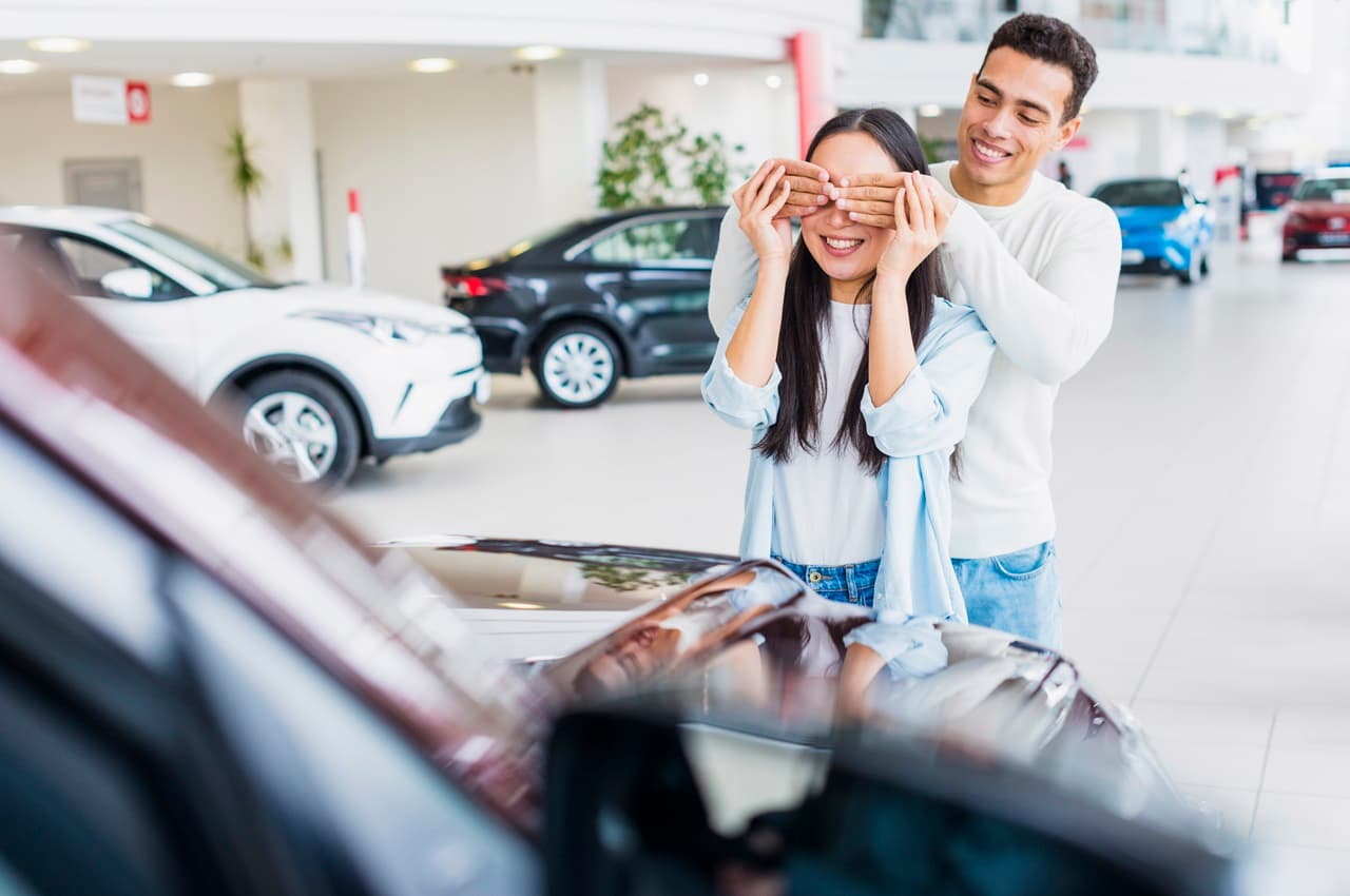 Homem sorridente tampando os olhos de uma mulher com as mãos para fazer surpresa de comprar carro na concessionária.