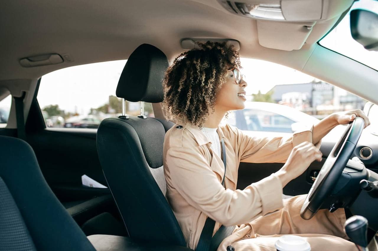 Mulher negra com expressão concentrada e terno bege claro dirigindo carro no trânsito.