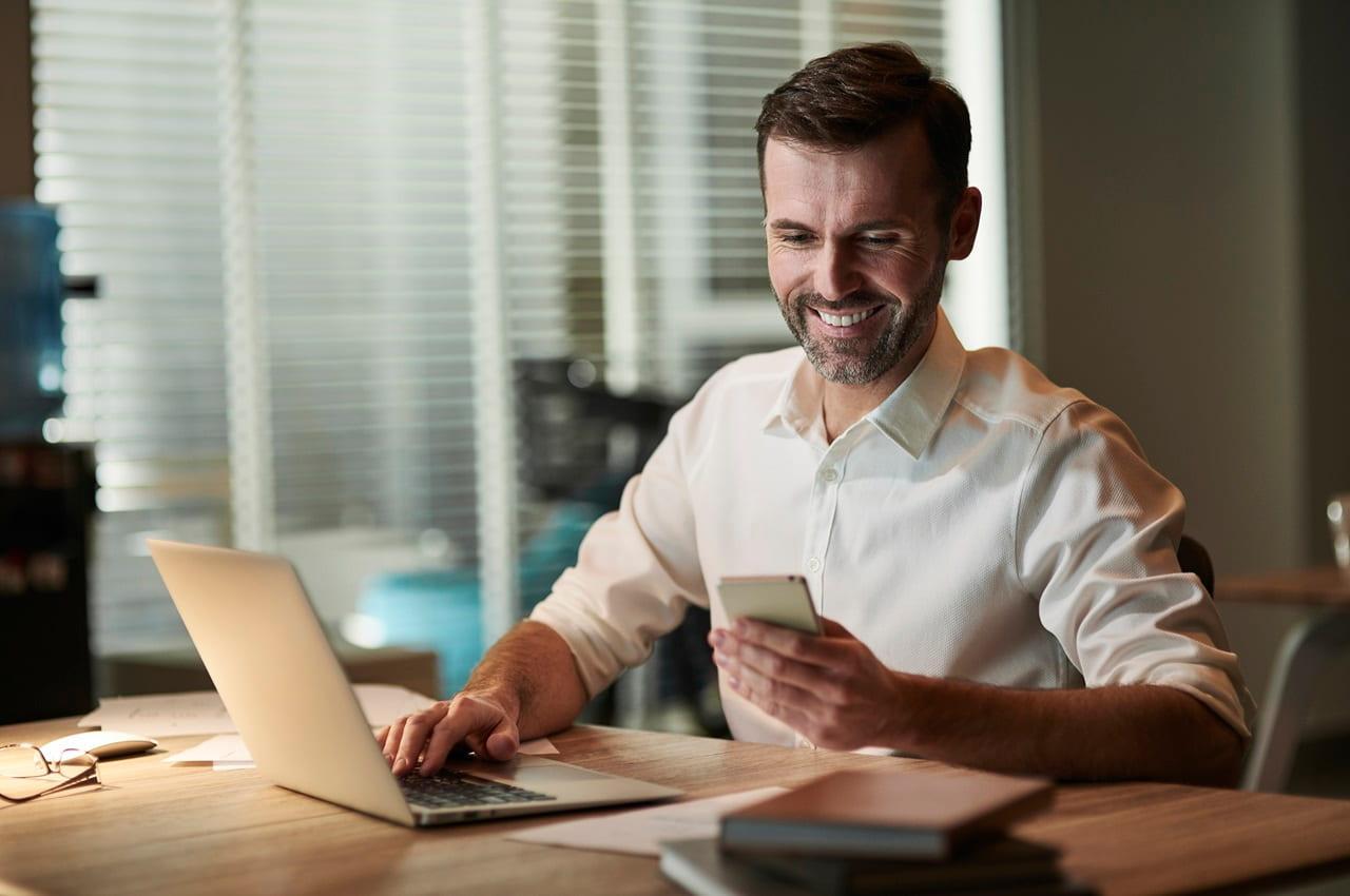 Homem sorridente usa celular e computador para consultar renavam de veículo. O homem veste camisa social de botões na cor branca.