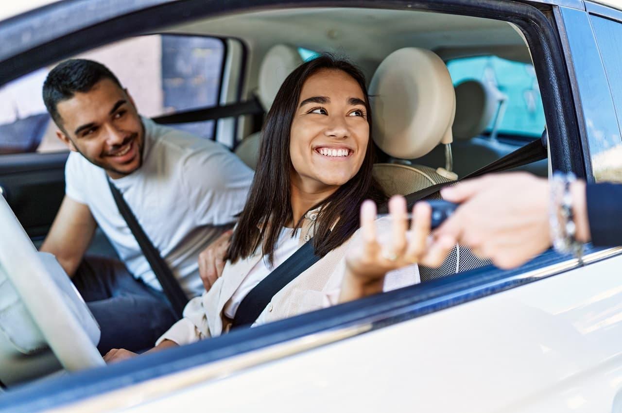 Mulher sorridente no banco de motorista de um veículo, recebendo chave de carro e, no banco de passageiro, um homem sorridente inclinado para frente observando a mulher.