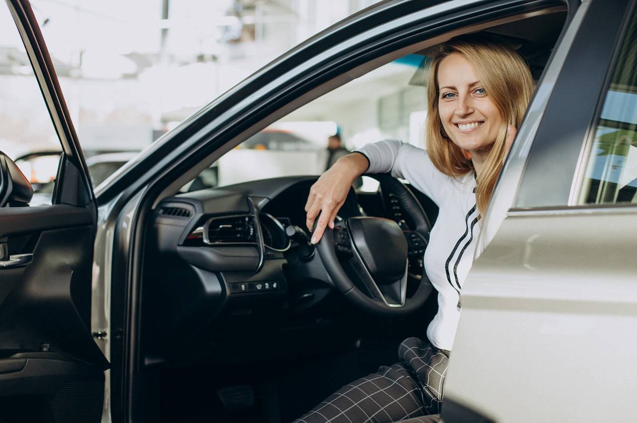 Mulher loira sorridente, vestindo blusa social branca e calça xadrez cinza. Ela está sentada no banco de motorista com a porta do veículo aberta para fazer test drive.