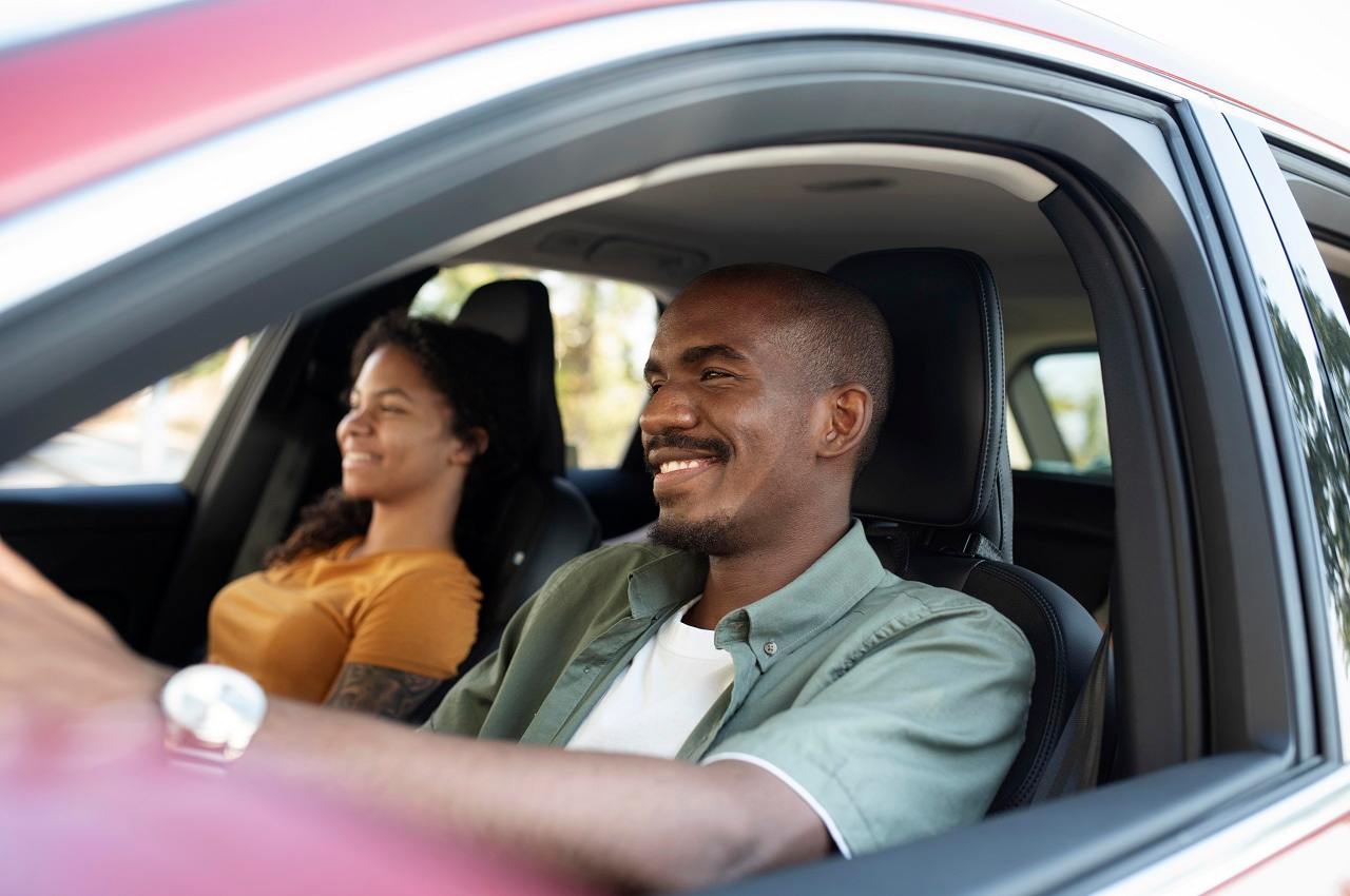 Homem sorridente dirigindo carro usado vermelho e no banco de passageiro ao lado tem uma mulher também sorridente.