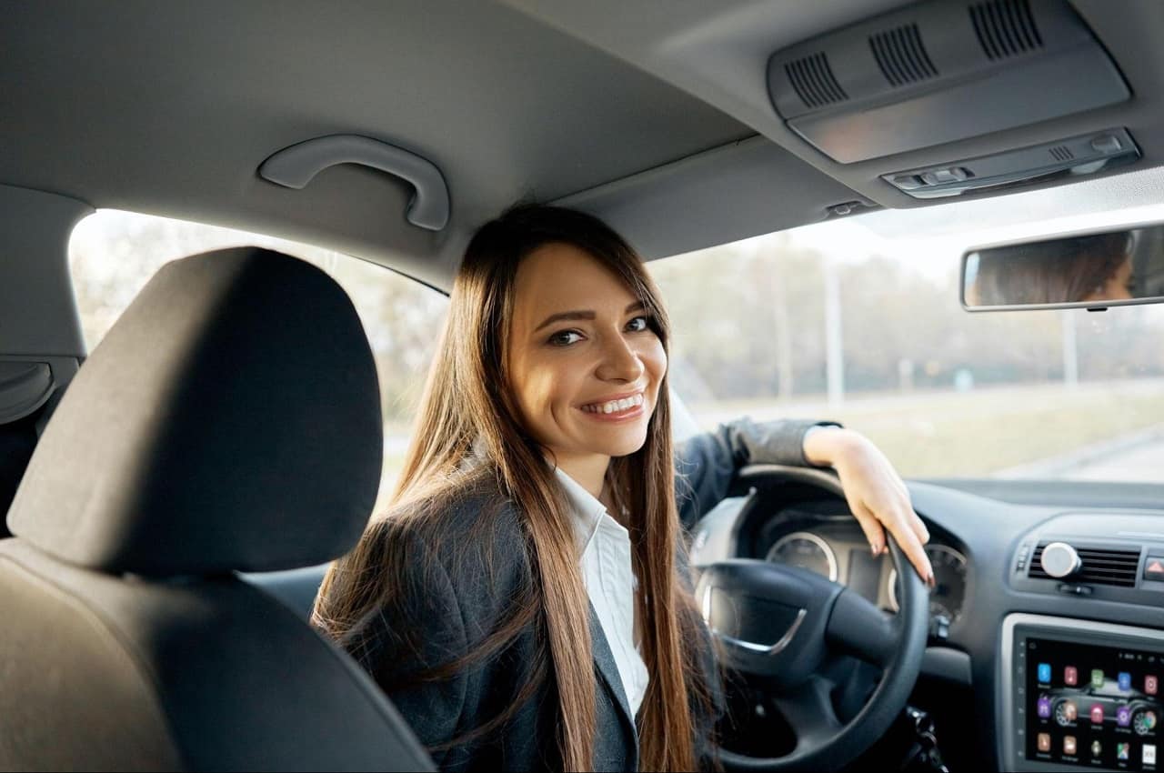 Mulher sorridente de longos cabelos castanhos e terno social, apoiada no volante de veículo e olhando para o banco passageiro.