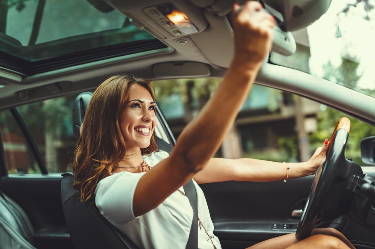 Mulher com expressão sorridente olhando para o espelho retrovisor do carro seminovo enquanto dirige.