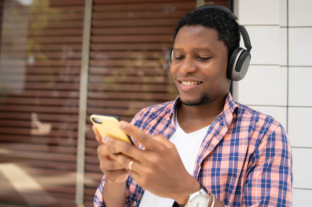 Homem negro sorridente com fones de ouvido bluetooth, blusa xadrez e digitando no celular para acessar whatsapp BV.