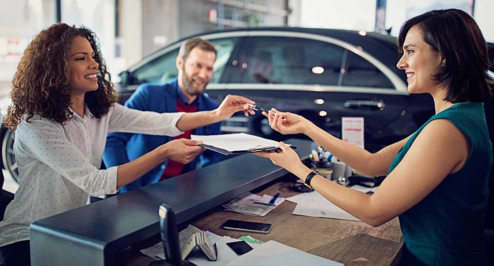 A imagem mostra um casal fechando negócio em uma concessionária. A funcionária está entregando o contrato para ser assinado.