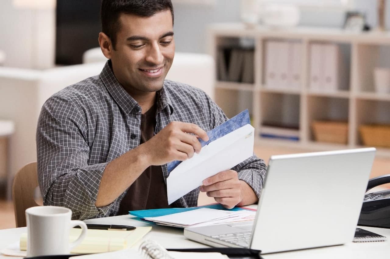 Homem sorridente abrindo carta com parcelas do financiamento e, em cima da mesa, tem um computador e anotações de contas.