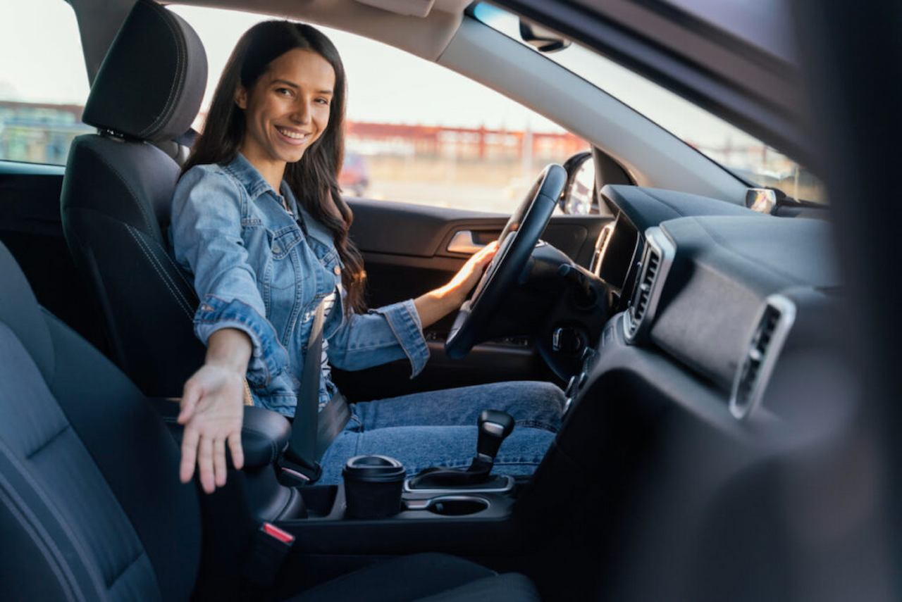 A imagem mostra uma mulher dirigindo seu carro. Ela está sorridente.