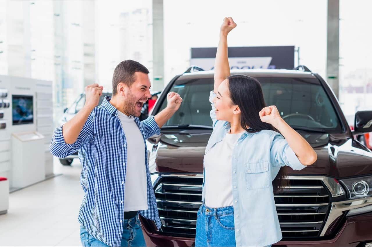 A imagem mostra um homem e uma mulher, a frente de um carro, dentro de uma concessionária, comemorando.