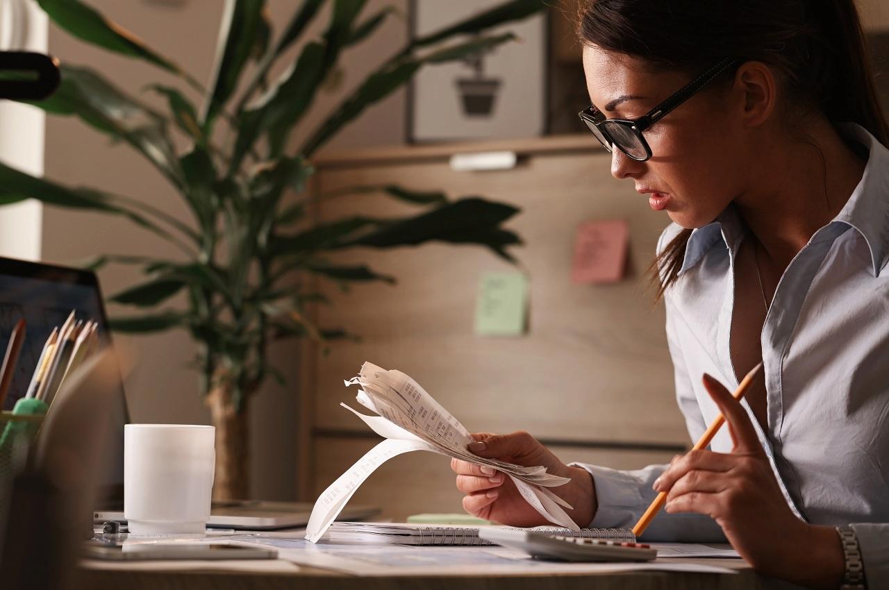 A imagem mostra uma mulher segurando algumas notas fiscais em uma mão, e na outra um lápis. Ao seu lado, um laptop e uma calculas