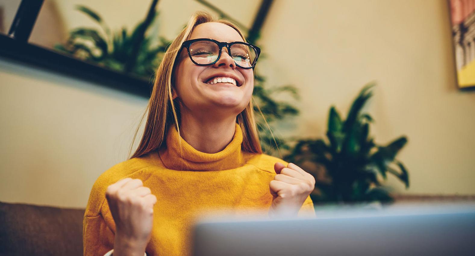 A imagem mostra uma mulher usando uma blusa amarela, usando seu laptop e comemorando.