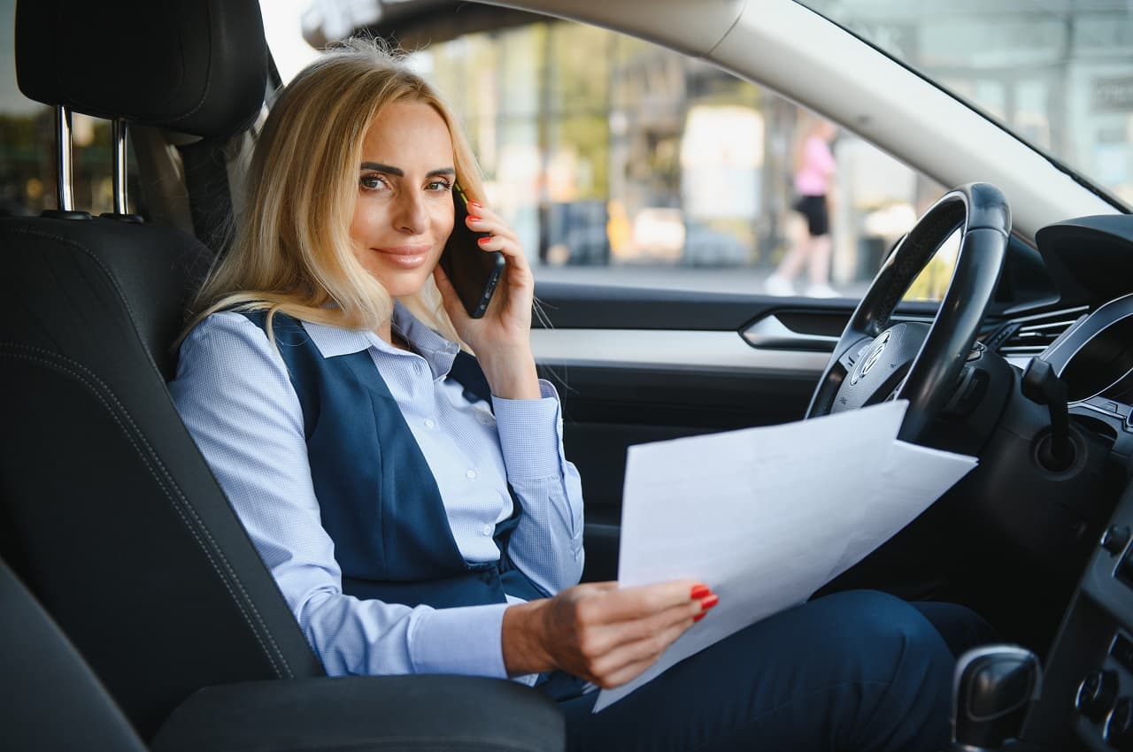 A imagem mostra uma mulher falando ao telefone dentro de seu carro. Ela está segurando alguns papéis.