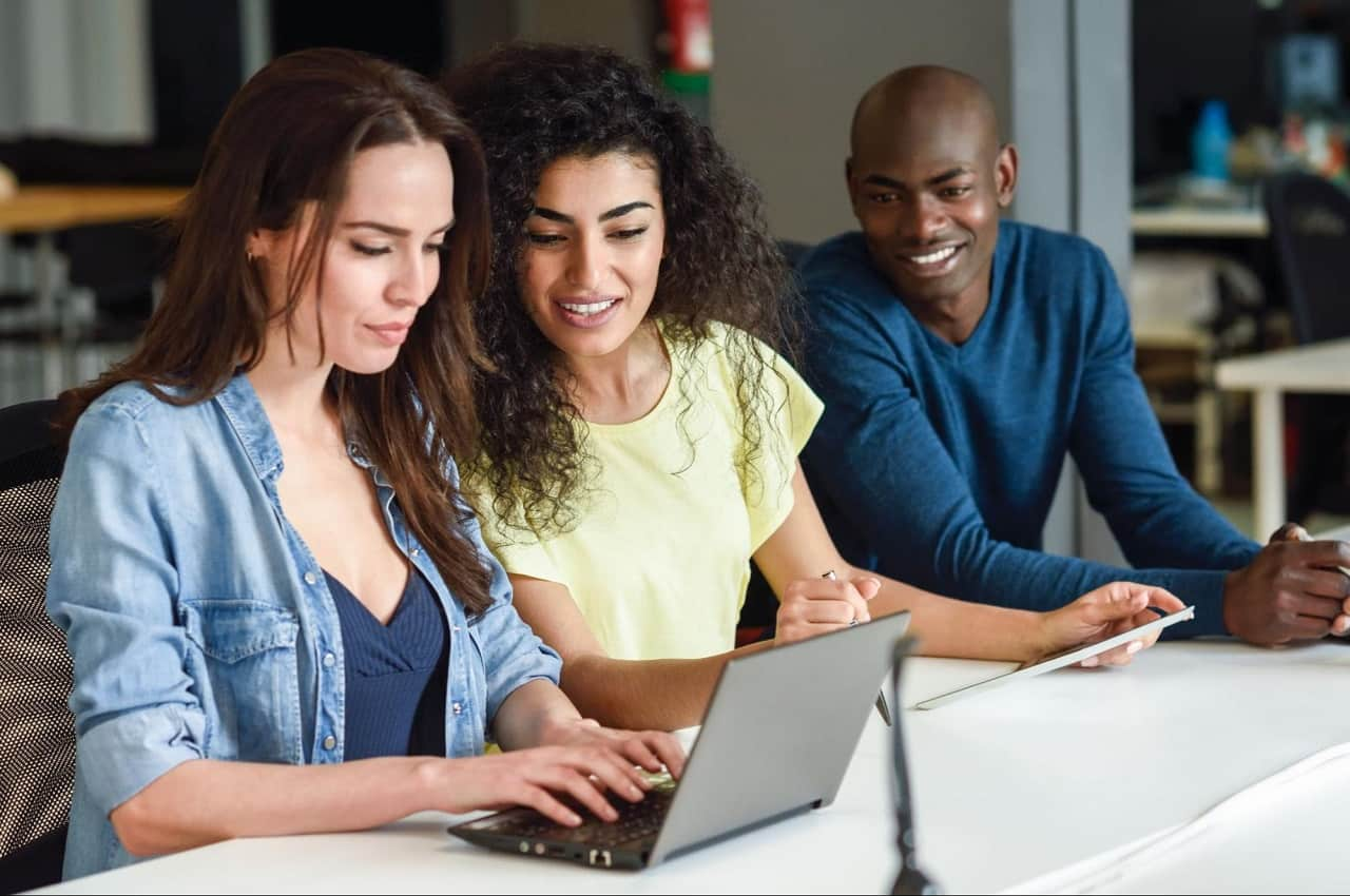 A imagem mostra duas mulheres e um homem sentados. Uma das mulheres está usando um laptop e todos estão olhando.