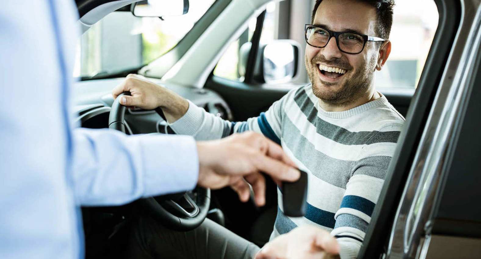 A imagem mostra um rapaz dentro de um carro em uma concessionária. Ele está no banco do motorista e sorridente.