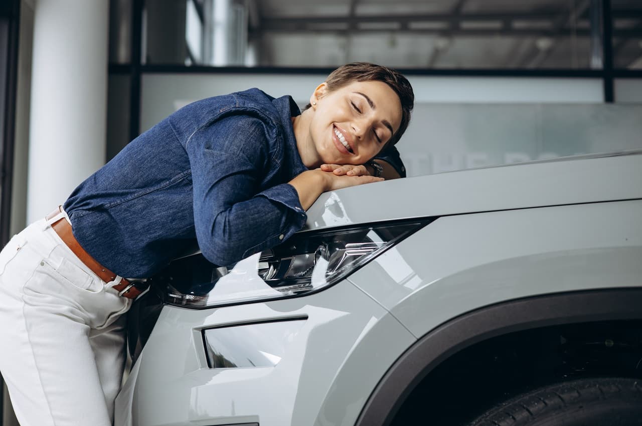 A imagem mostra uma mulher sorridente, deitada no capô de um carro.