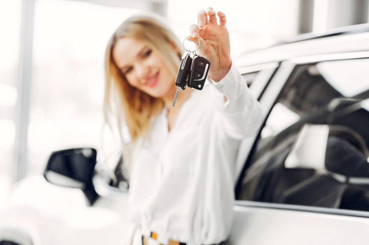 A foto mostra uma mulher loira, encostada em um carro branco, segurando suas chaves.