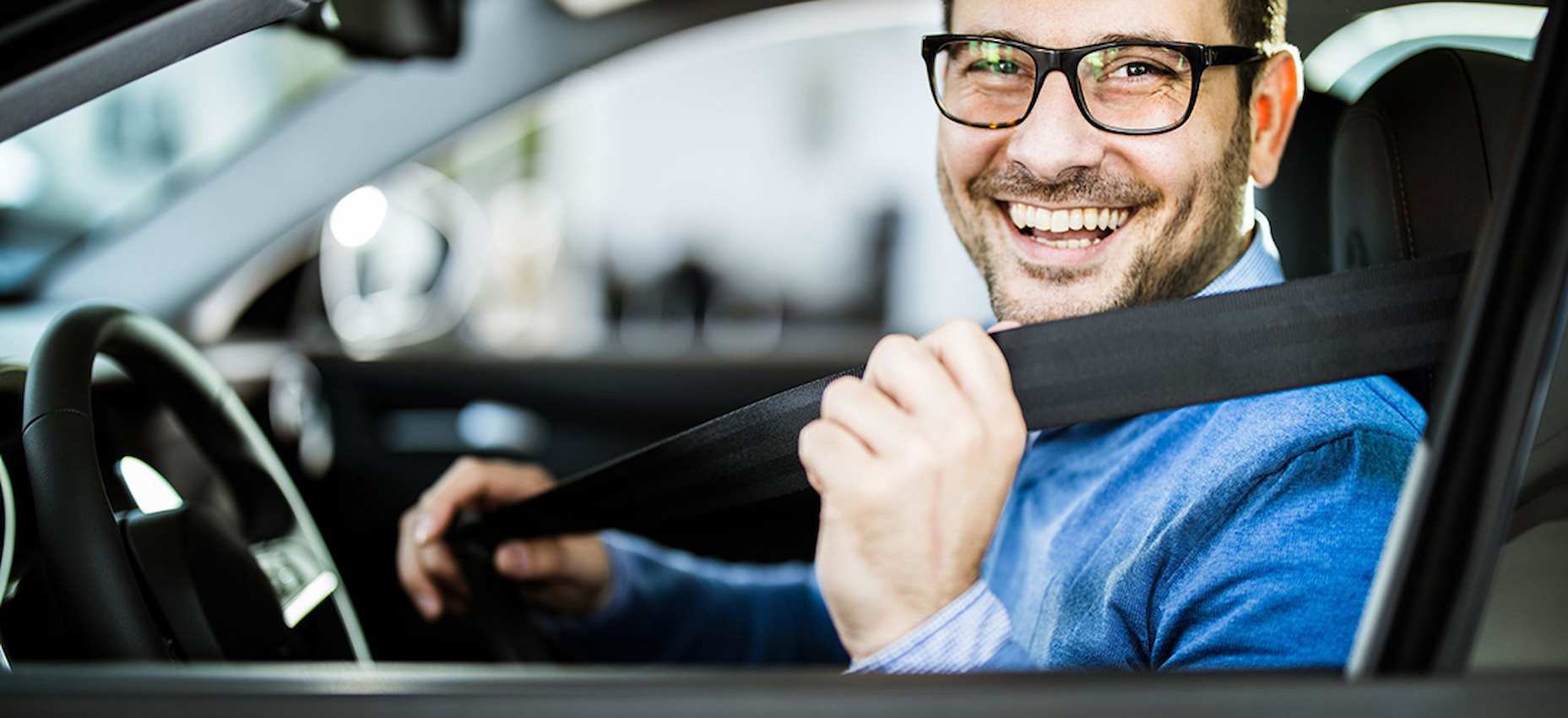 A foto mostra um rapaz, dentro de seu carro, sorrindo e colocando o cinto de segurança.