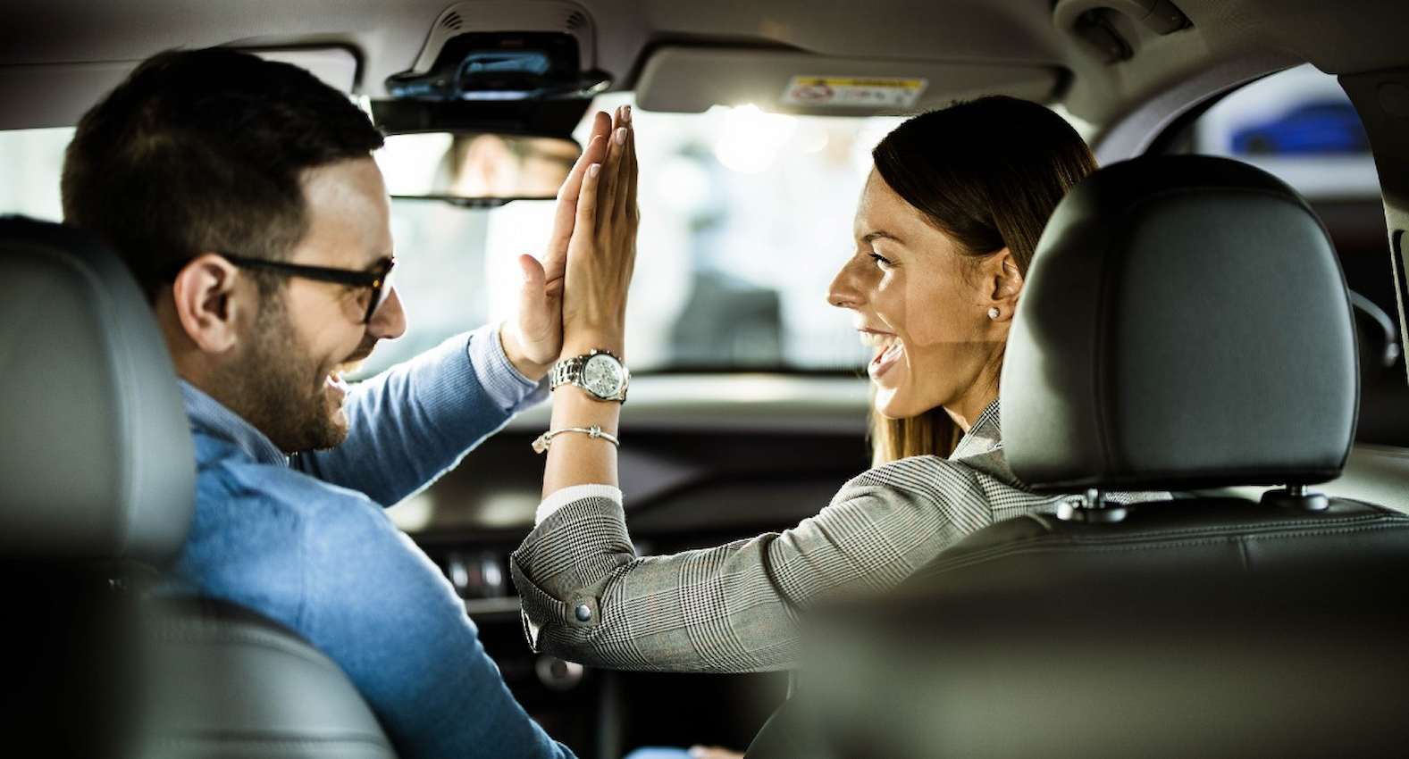 A imagem mostra um homem e uma mulher sentados no banco da frente de um carro e tocando as mãos. Eles estão sorridentes.