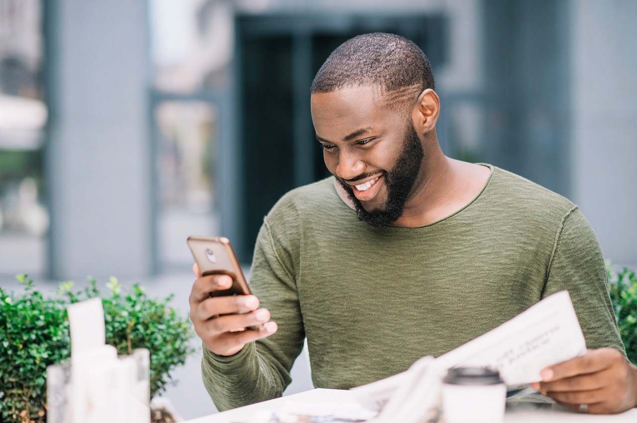 A imagem retrata um homem, segurando alguns papéis e sorrindo para seu celular. Ele está em uma mesa ao ar livre.