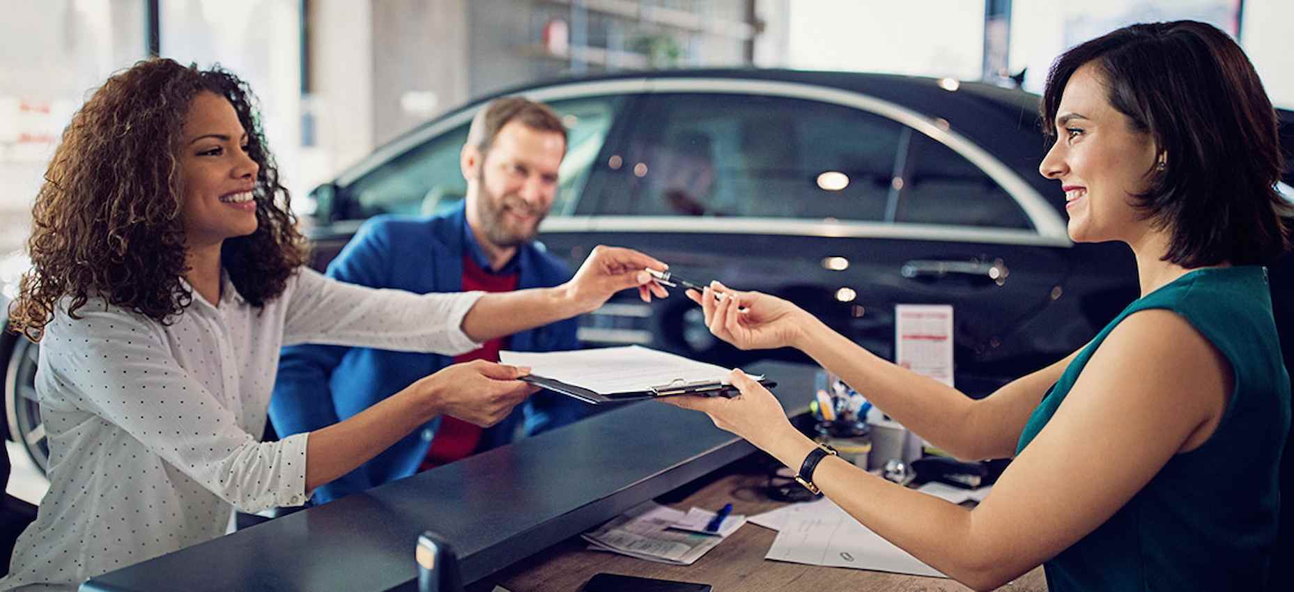 A imagem mostra uma mulher fechando um negócio em uma concessionária. A funcionária da concessionária está entregando o contrato para ser assinado.