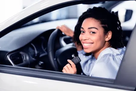 Imagem de uma mulher sorrindo dentro de um carro, mostrando a chave dele com uma mão e, com a outra, segurando o volante.