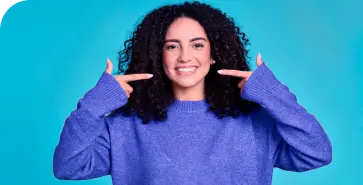 A imagem mostra uma mulher branca. Ela veste uma camisa azul, com seus dedos apontados para seus dentes e em um fundo azul.