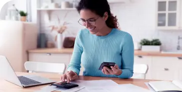 Mulher branca de óculos de grau e cabelos escuros. Ela veste blusa azul e usa celular, laptop e calculadora em sua cozinha.