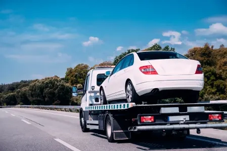 Carro branco sendo transportado por um caminhão de reboque na estrada. O dia está ensolarado e, ao fundo, há muitas árvores.