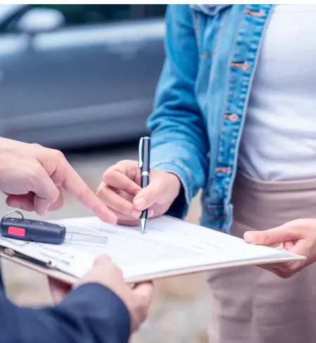 Duas pessoas assinam contrato. A primeira aponta para um formulário anexo a uma prancheta e uma chave de carro, a outra assina.