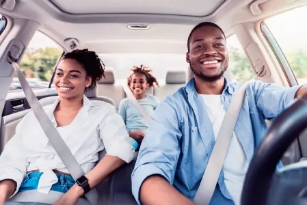 Uma familia de três pessoas viaja de carro. Um homem negro, de camisa azul, dirige, levando uma mulher e uma criança.