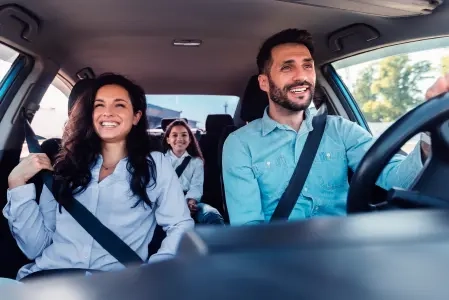 A imagem mostra uma família sorridente de três pessoas em um carro, usando cinto de segurança. Ambiente ensolarado.