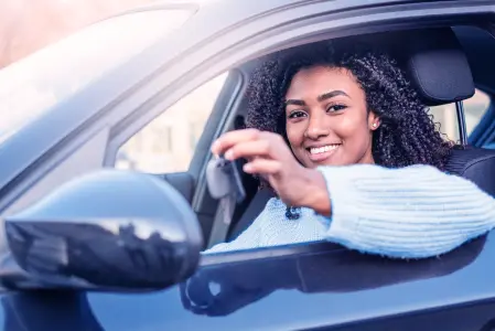 Mulher negra em carro preto, mostrando chave, vestindo blusa azul clara, sorrindo para câmera.