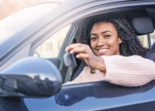 Mulher negra em carro preto, mostrando chave, vestindo blusa azul clara, sorrindo para câmera.