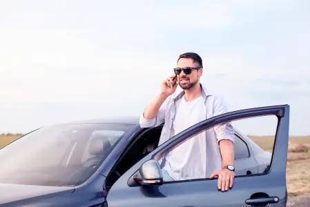 Homem de barba e óculos escuros fala ao celular perto de carro, vestindo camisa azul e camiseta branca, mão apoiada na porta.