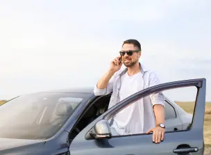 A foto mostra um homem de barba e óculos escuros falando ao celular. Ele está vestindo uma camisa azul-clara e uma camiseta branca. Ele está do lado de fora do carro, com uma mão apoiada na porta do veículo.