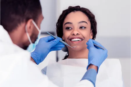 Mulher sorrindo em um consultório de dentista enquanto é examinada por um rapaz com instrumentos odontológicos.