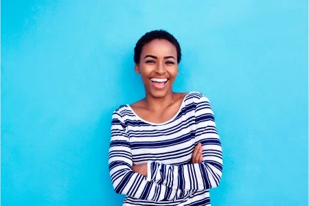 Mulher de cabelos curtos, usando camisa listrada azul e branca, com os braços cruzados e sorrindo para a câmera.