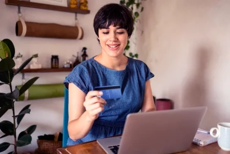 A imagem mostra uma mulher branca, de cabelos curtos, sentada em frente a um laptop aberto, segurando um cartão de crédito.