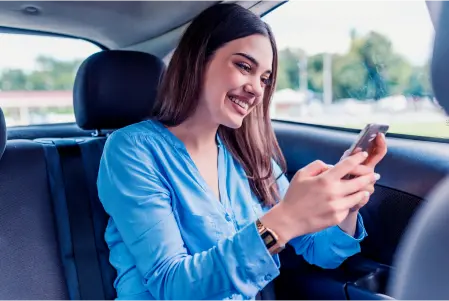 Mulher de longos cabelos escuros sorrindo, segurando um celular com as duas mãos, sentada no banco do motorista de um carro.