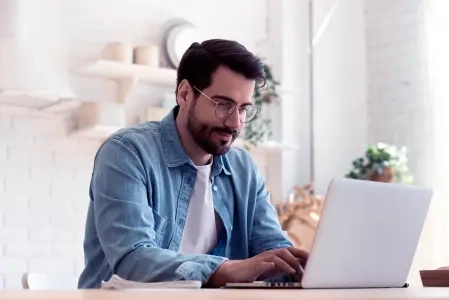 A imagem mostra um homem de óculos de grau, vestindo uma camisa branca e uma jaqueta jeans azul, sentado e usando um notebook