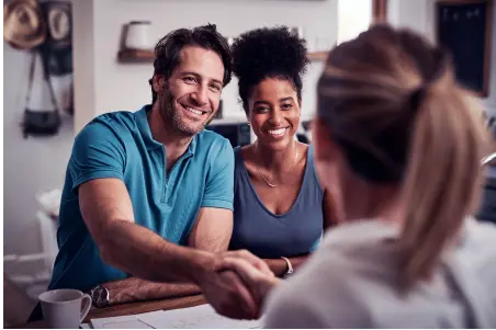 Um casal sentado à mesa, sorrindo e segurando as mãos de outra mulher à sua frente, como se estivessem fechando um negócio.