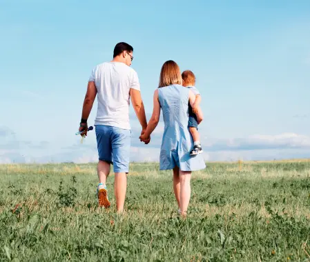 Um homem, uma mulher, um bebê de colo e um cachorro caminham juntos em um campo ensolarado, estão de costas com as mãos dadas