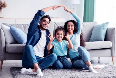 Imagem com dois adultos e uma criança sentados no chão da sala formando um telhado de casa com os braços na frente de um sofá