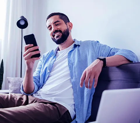 Homem branco sentado em um sofá, sorrindo, olhando para o celular que está em sua mão