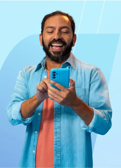 Um homem de barba sorri ao usar o celular. Ele veste camisa jeans azul clara e camiseta coral.