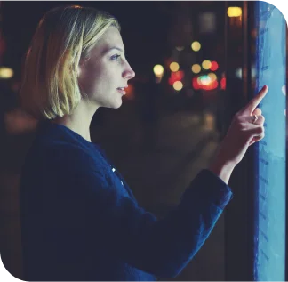 Em uma rua, à noite, há uma tela touchscreen de lado e uma mulher loira de cabelo curto e blusa azul marinho interage com ela com seu dedo indicador. 