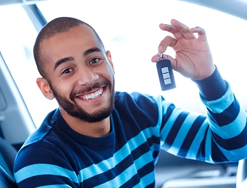 A imagem mostra um rapaz negro, jovem, de barba, no interior de um carro. Ele está do lado do motorista e segura a chave do carro com a mão esquerda enquanto olha pra frente e sorri. Ele está vestindo uma blusa de manga longa azul escura com listras em azul claro.