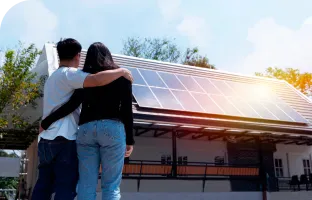 Casa com telhado de painéis solares, cercada por árvores, com um casal se abraçando do lado de fora e olhando para ela.